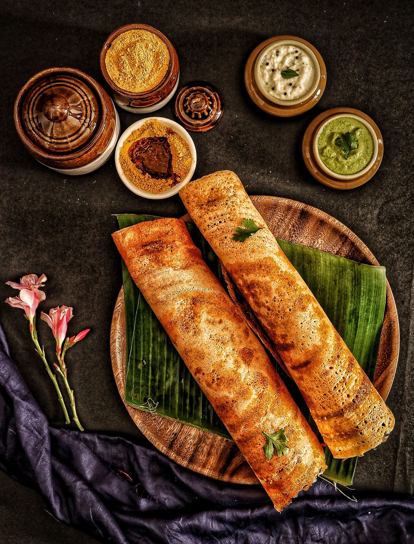 Crispy dosa rolls on a banana leaf with chutneys and spice powders.