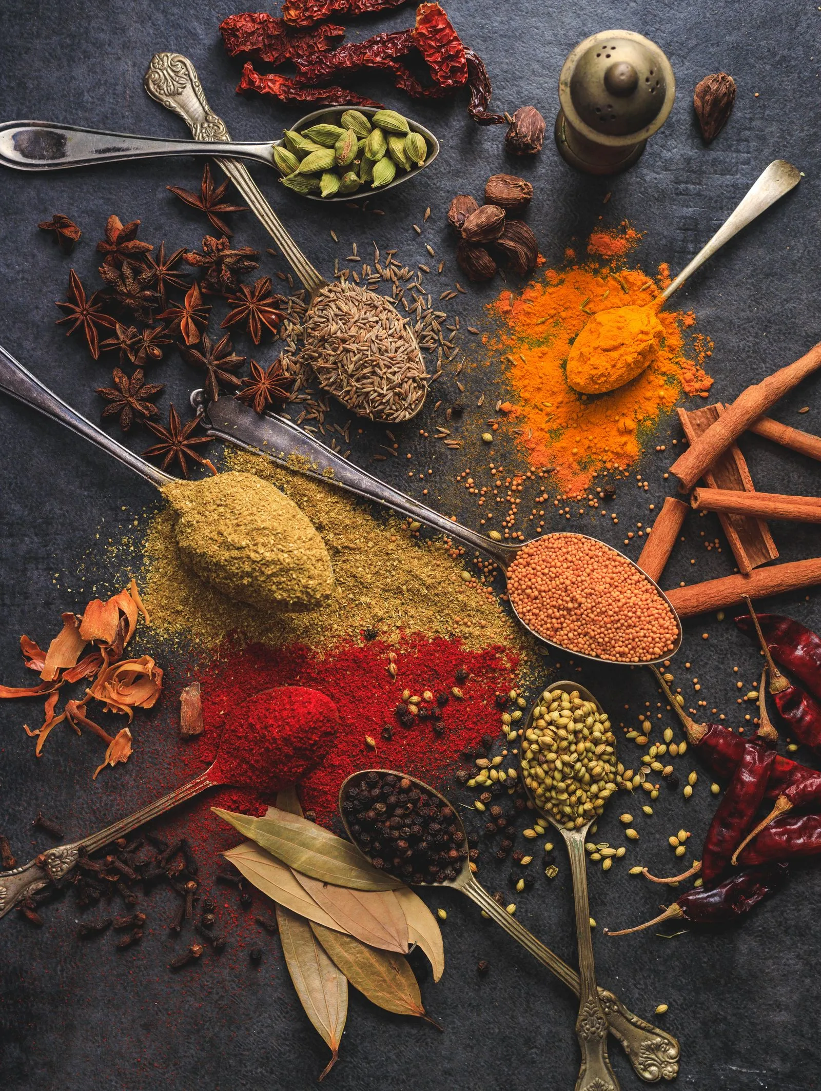Spoons of turmeric, chilli, mustard, cardamom, cinnamon and other spices on a dark surface.