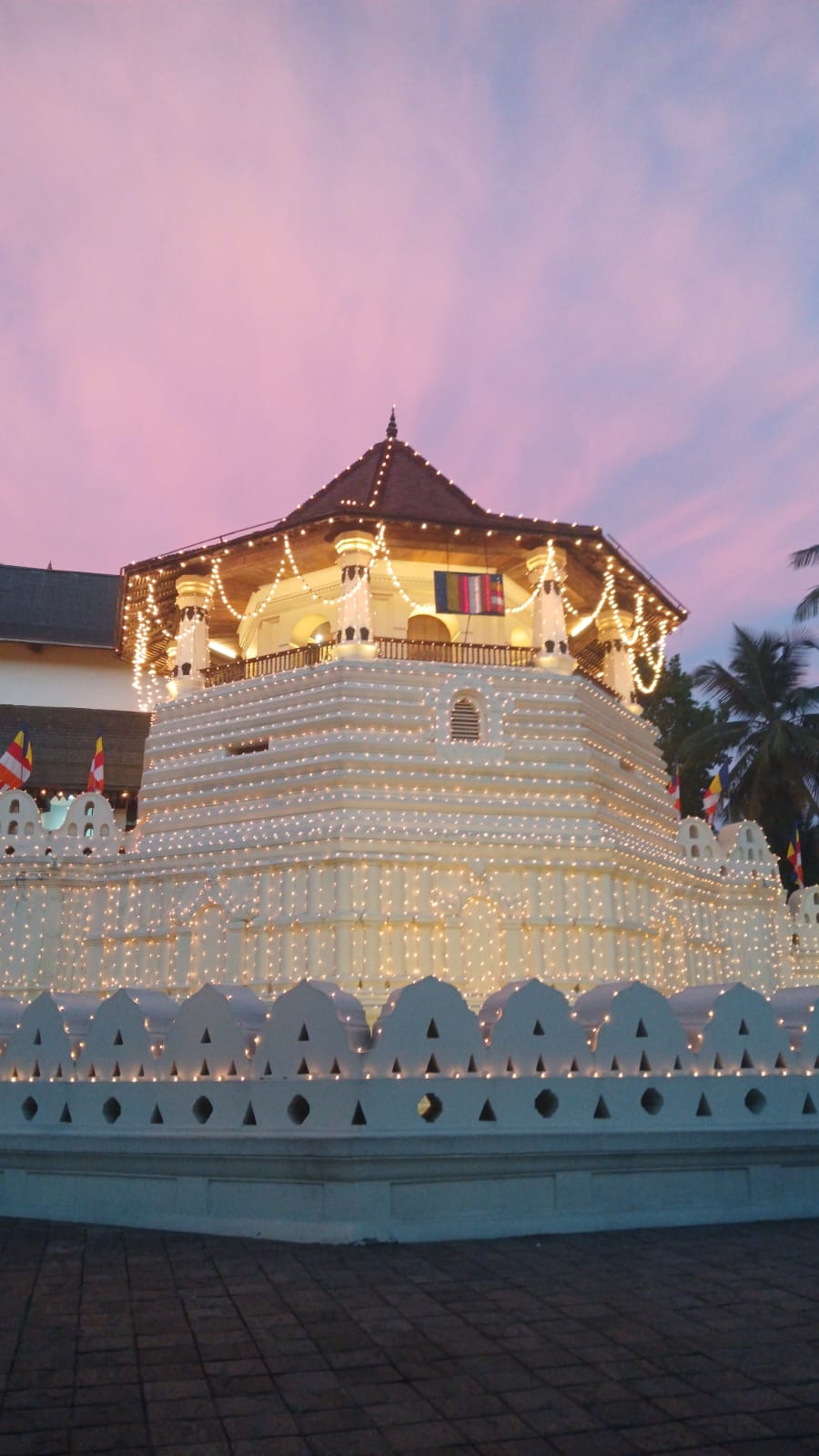 The illuminated octagonal pavilion of the Temple of the Sacred Tooth Relic beneath a pink evening sky.
