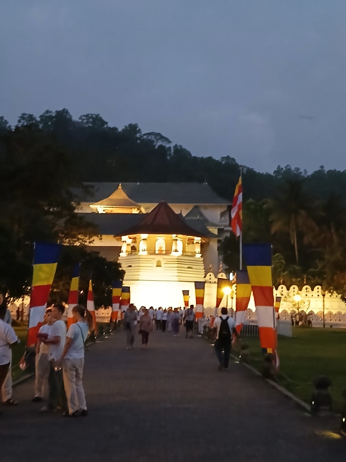 Visitors walking toward the Temple of the Sacred Tooth Relic between Buddhist flags at dusk.